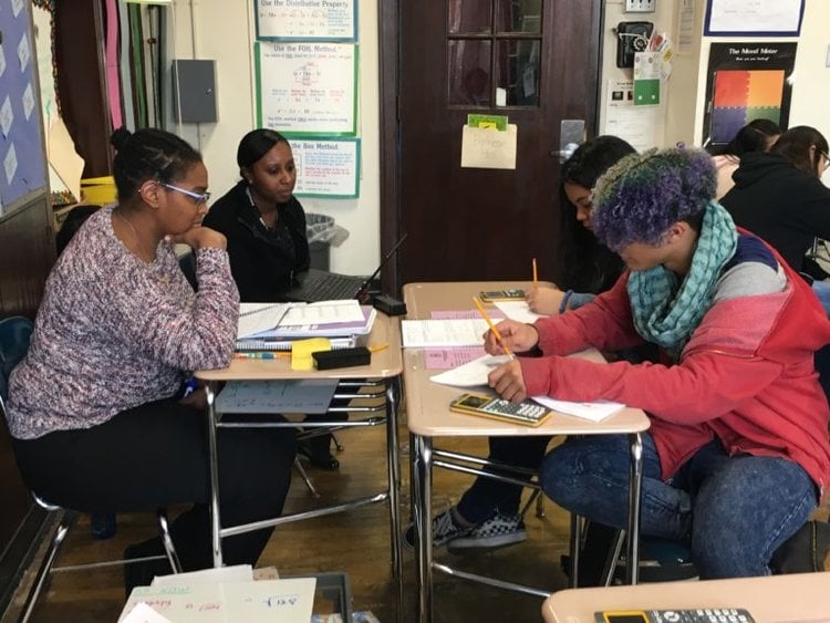 Two tutors sitting across from 2 students who are holding pencils and calculators working on a math problem on their desks in a classroom 